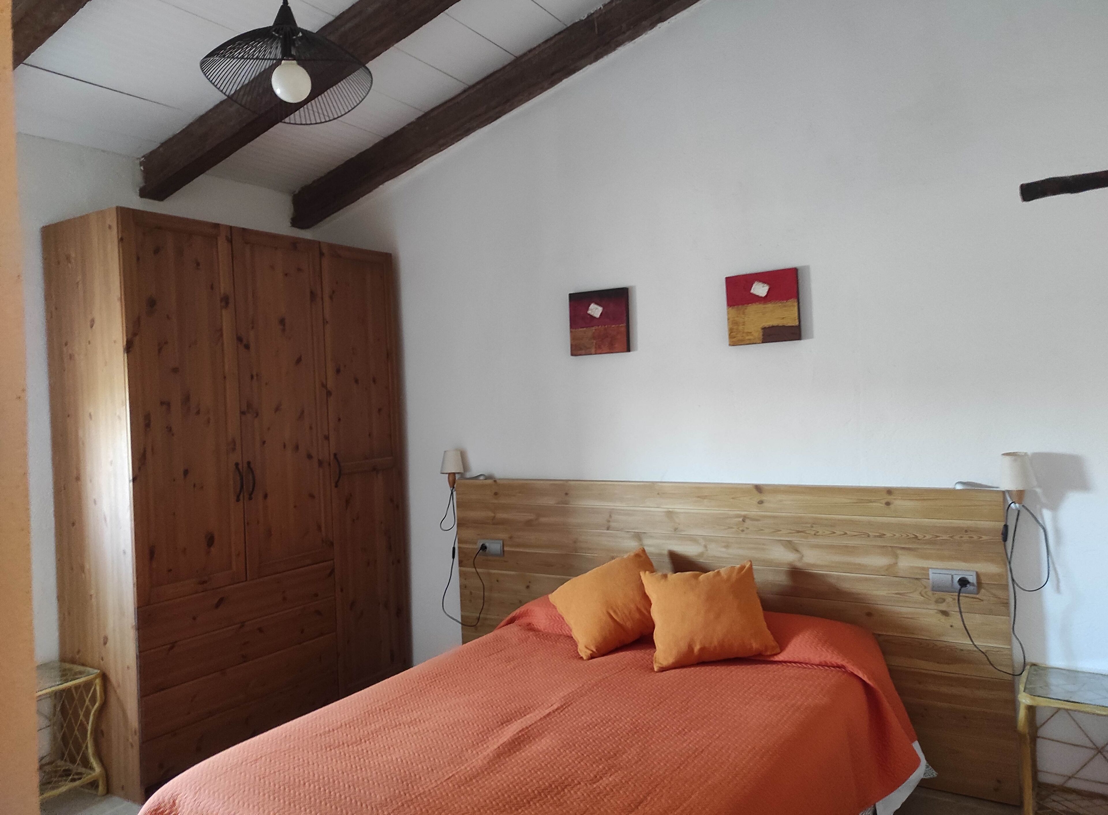 A bedroom with a wooden headboard, a bed with an orange blanket, two pillows, a wooden wardrobe, and two wall-mounted paintings.