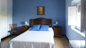 Une chambre à coucher avec un lit, deux tables de chevet, une chaise, une commode en bois, un tableau au mur et une fenêtre avec des stores.