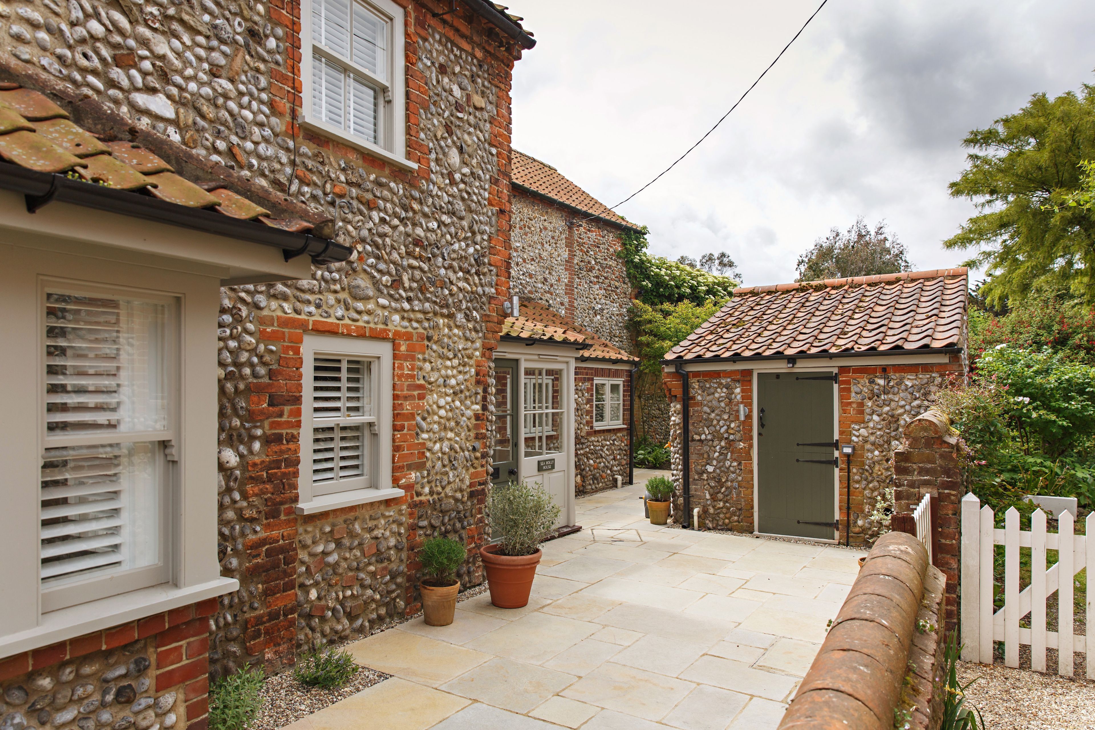 A stone building with a tiled roof, a white picket fence, and a small garden area with potted plants.