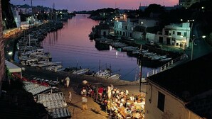 Un puerto al atardecer con barcos amarrados, gente cerca del paseo marítimo y edificios a lo largo de la costa.