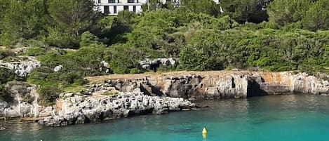 Un hotel frente al mar con costa rocosa, agua turquesa cristalina y exuberante vegetación.