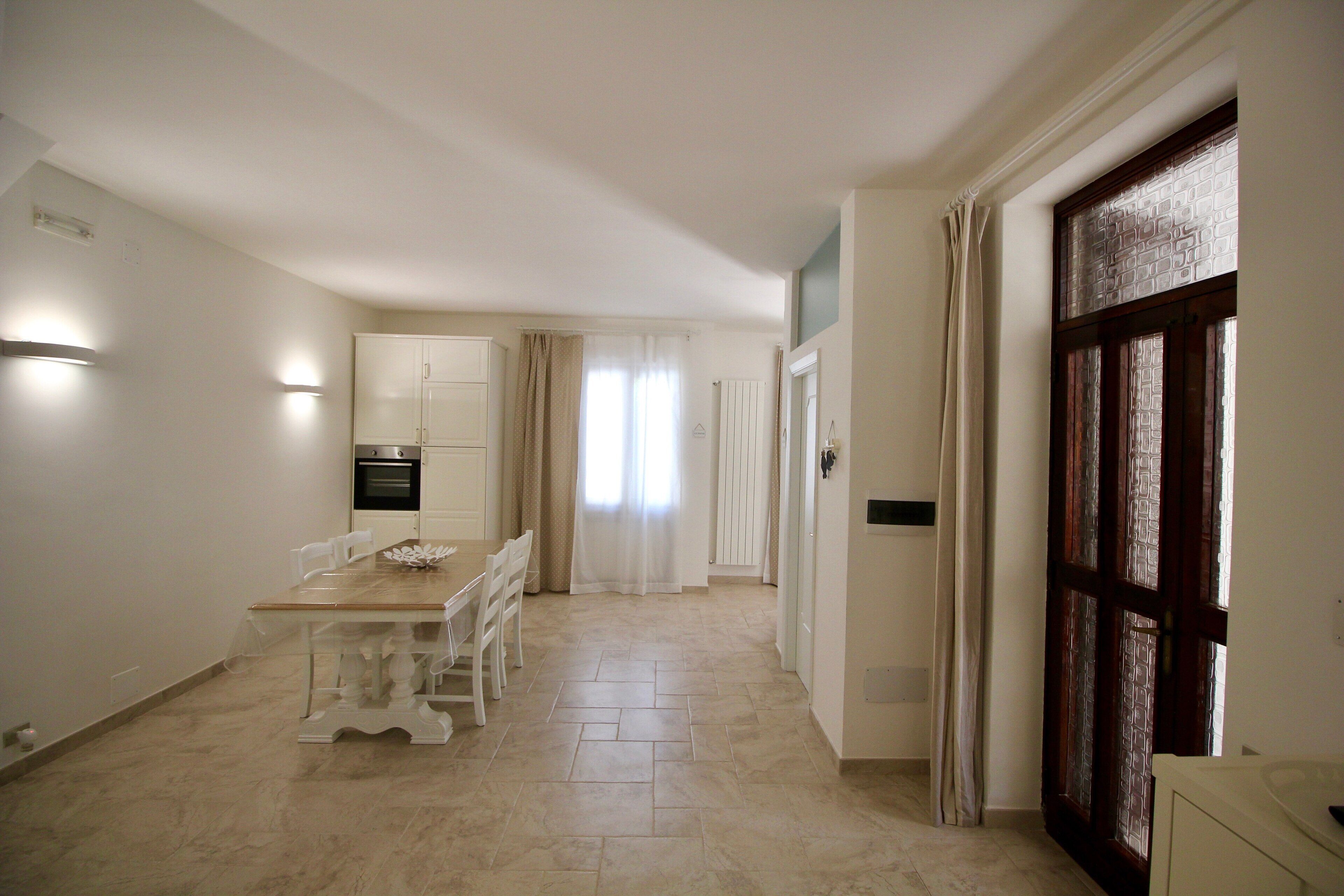 A spacious kitchen with a dining area, a large window with curtains, and a wooden door.