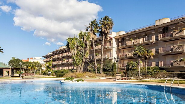 Ein Pool mit klar blauem Wasser, umgeben von einer begrünten Anlage mit Palmen und einem mehrstöckigen Gebäude mit Balkonen.