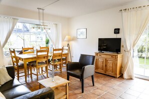 A living room with a sofa, a dining table with chairs, a TV on a wooden cabinet, and a window with curtains.