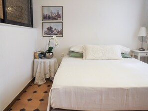 A neatly arranged bedroom with a bed, a small bedside table with a lamp, a wall-mounted shelf with books and a potted plant, and two framed pictures on the wall.