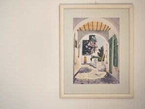 A framed artwork depicting a narrow alleyway with a stone pathway, an archway, and a tree with pink flowers. - Haraki beach house. Beautiful, family-friendly, perfect for relaxing holiday (Rhodes)