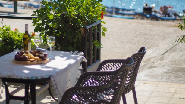 Una mesa con una botella de vino y una copa, rodeada de sillas, con una piscina y sombrillas al fondo.