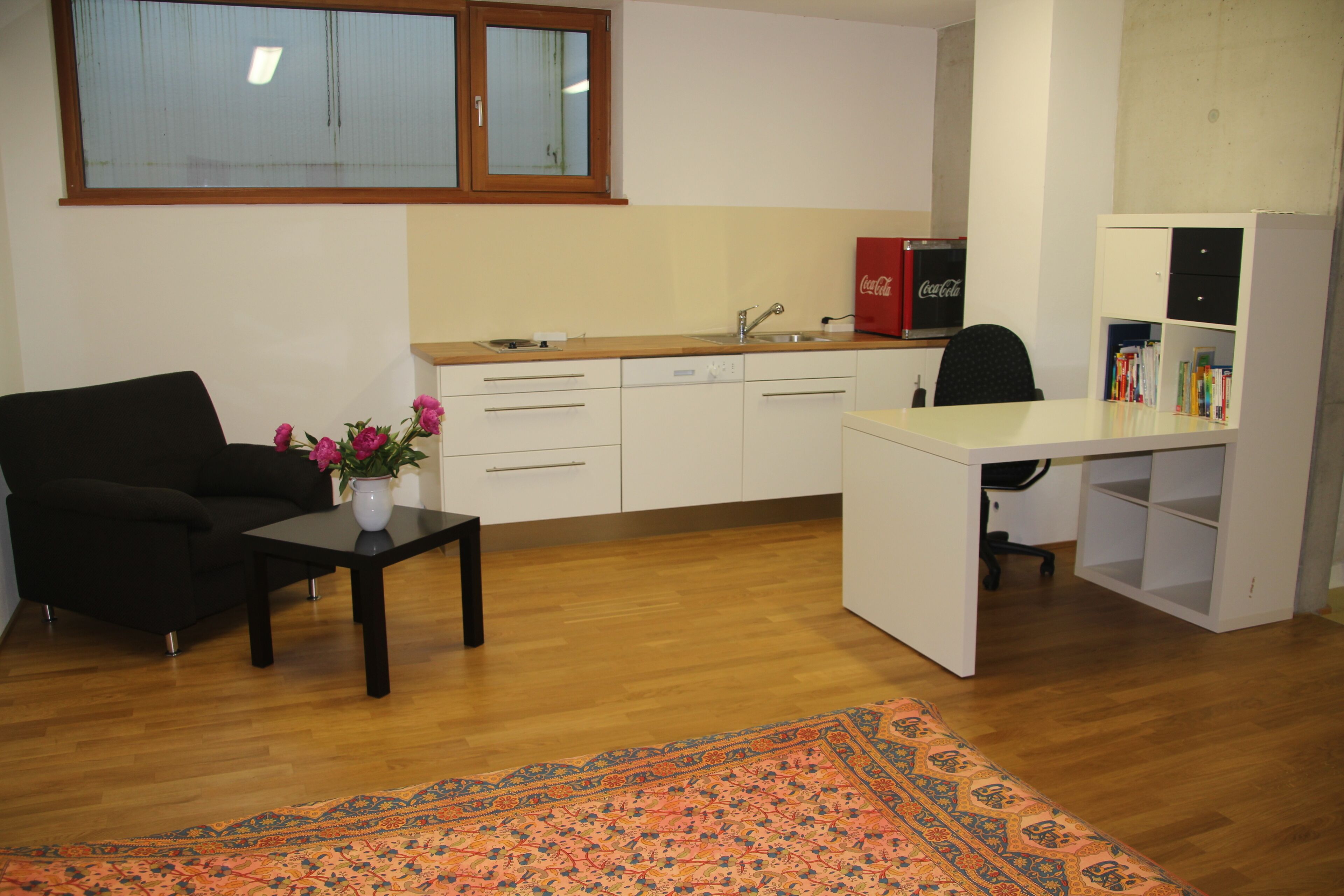 Ein kompakter Wohnbereich mit Sofa, einem kleinen Tisch mit Vase, einer Küchenzeile mit Spüle und Schränken sowie einem Schreibtisch mit Stuhl.