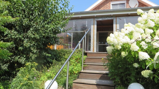Una casa moderna con una escalera de madera que conduce a una puerta de vidrio, rodeada de abundante vegetación y flores blancas.