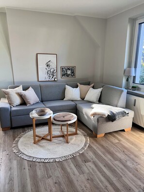 Un salon moderne avec un canapé d’angle gris, deux tables d’appoint, un tapis rond et des œuvres d’art encadrées accrochées au mur.