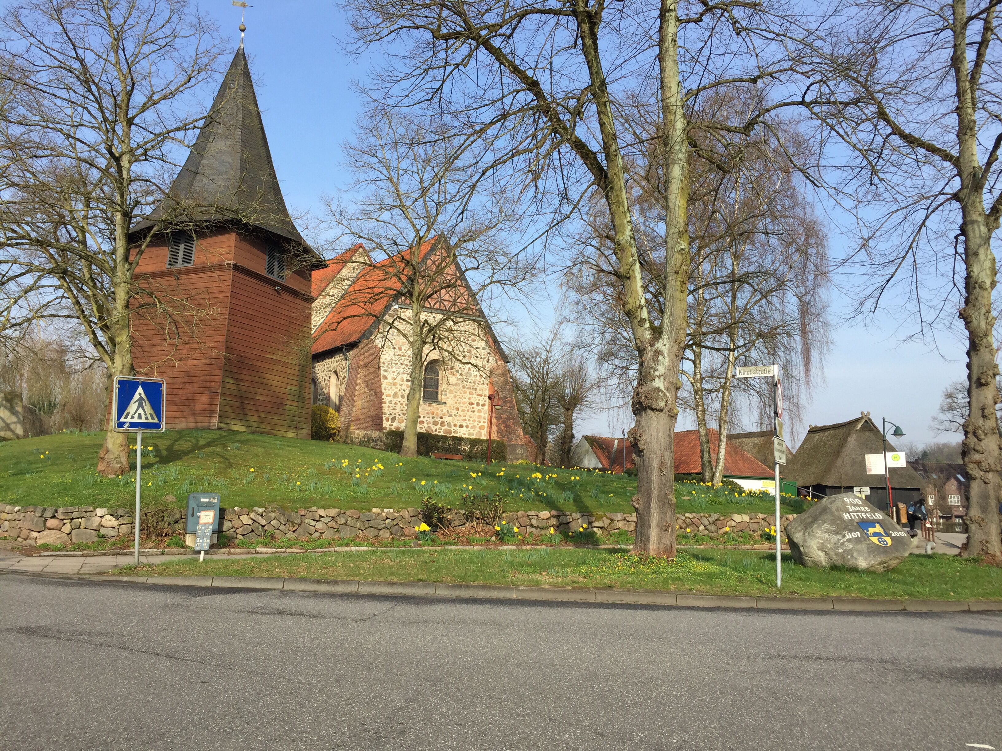A church with a stone facade and a red roof, a pedestrian crossing sign, and a stone wall with a rock inscribed with names.