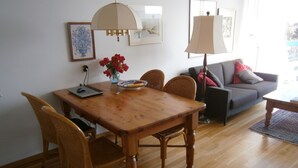 A dining area with a wooden table, chairs, and a vase of red flowers. There is a grey sofa and a lamp in the background.