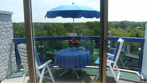 A balcony with a table and chairs, a blue umbrella, and a view of greenery.