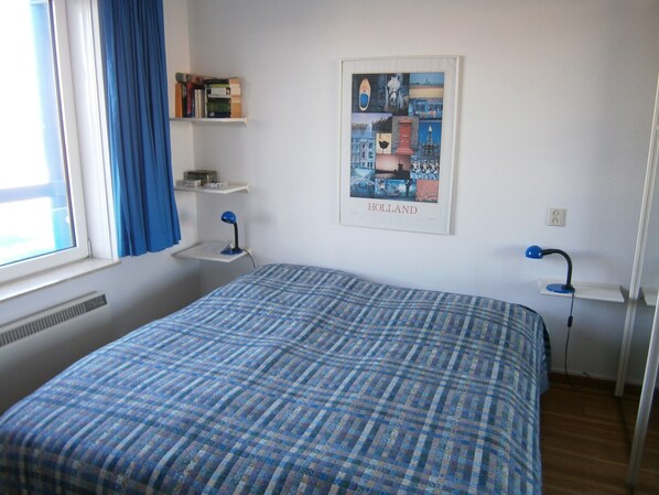 Une chambre avec un lit, un bureau avec une lampe, une fenêtre avec des rideaux, une étagère murale avec des livres et une affiche encadrée représentant un paysage urbain.