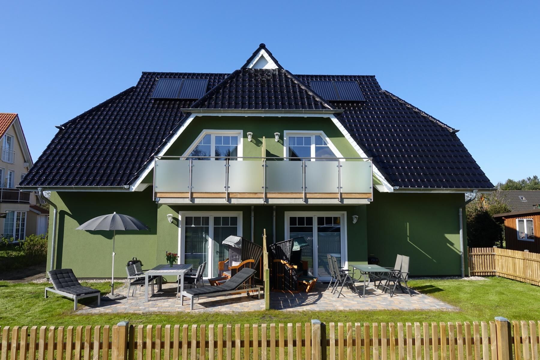 A two-story house with a green exterior, a dark tiled roof, and a balcony with glass railings.