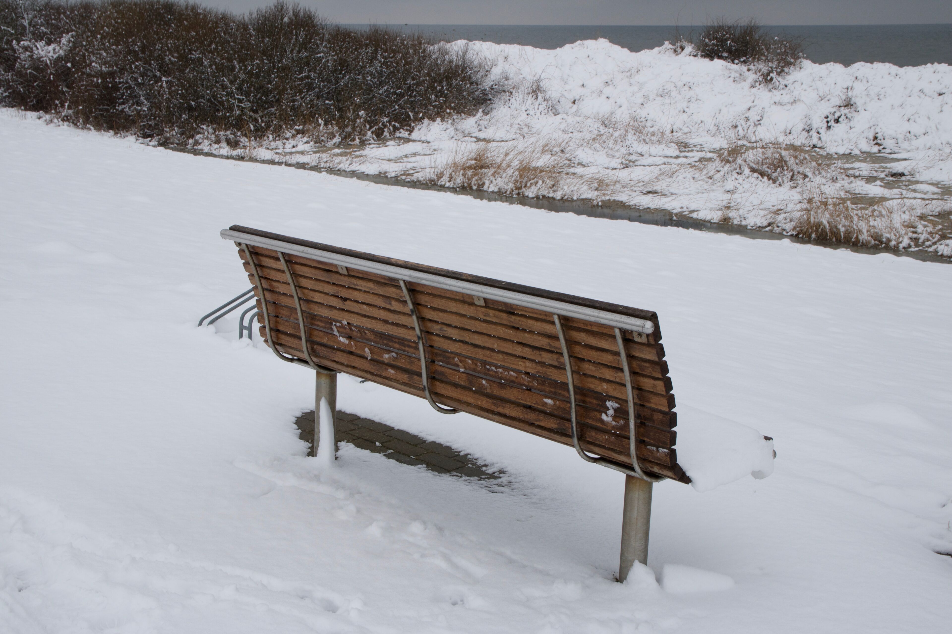 A wooden bench in a snowy area with a view of the sea in the background.