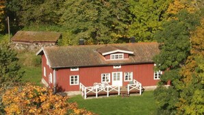 Una casa de madera roja con un porche blanco y una pequeña terraza, rodeada de abundante vegetación y árboles.