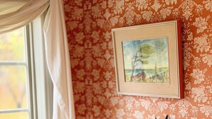 Una habitación con una silla blanca, una mesa de madera, un radiador y un papel tapiz con motivos florales.