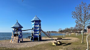Ein Spielplatz mit zwei blau-weiß gestreiften Türmen, einer Schaukel und einer Bank in der Nähe eines Gewässers.