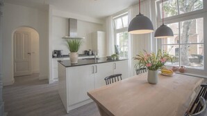 Una cocina moderna con armarios blancos, una isla central, iluminación colgante y vistas amplias a través de grandes ventanales.