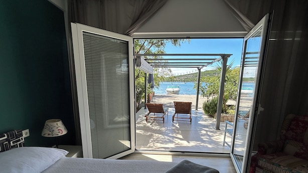 A bedroom with a bed, a bedside table, and a view of a beach with a boat and lounge chairs.