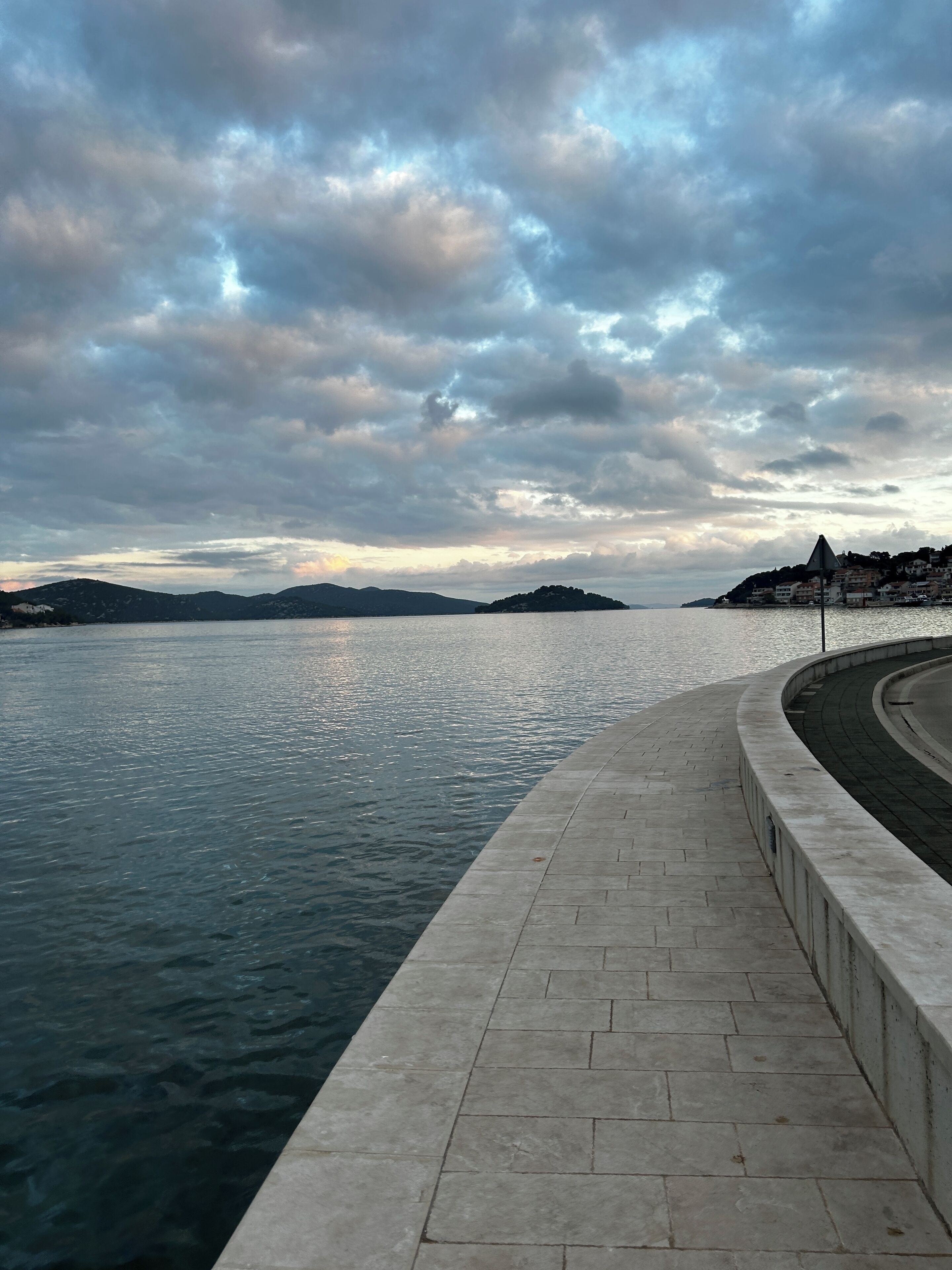 A waterfront promenade with a paved walkway and a view of the water and distant hills.