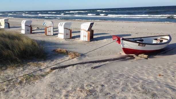 A beach with white wicker chairs lined up, a small boat with 'KR 02' on it, and the ocean in the background.