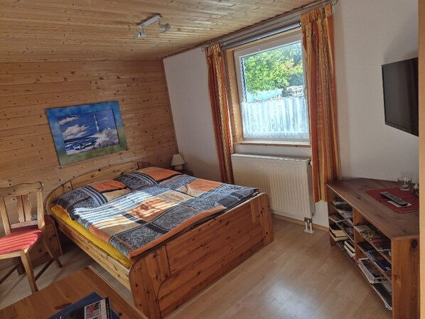 A cozy room with a wooden bed, a window with curtains, a painting on the wall, and a TV on the wall. - Apartment i haven, Germany (Steinbergkirche)