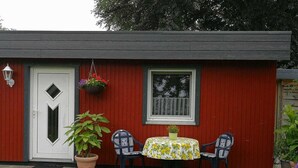 A small red building with a white door, a window, and a round table with chairs. There are potted plants and a hanging flower basket.