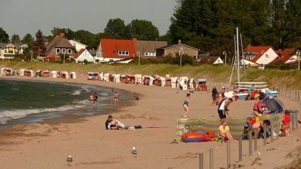 Une plage avec des maisons en arrière-plan, un rivage sablonneux, ainsi que des personnes et des bateaux.