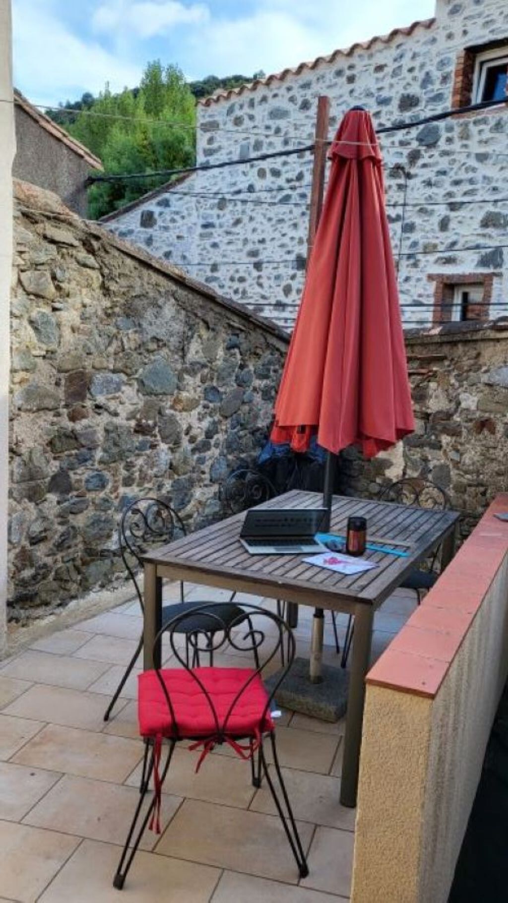 Un pati a l'aire lliure amb una taula i cadires, un parasol vermell i un mur de pedra al fons.