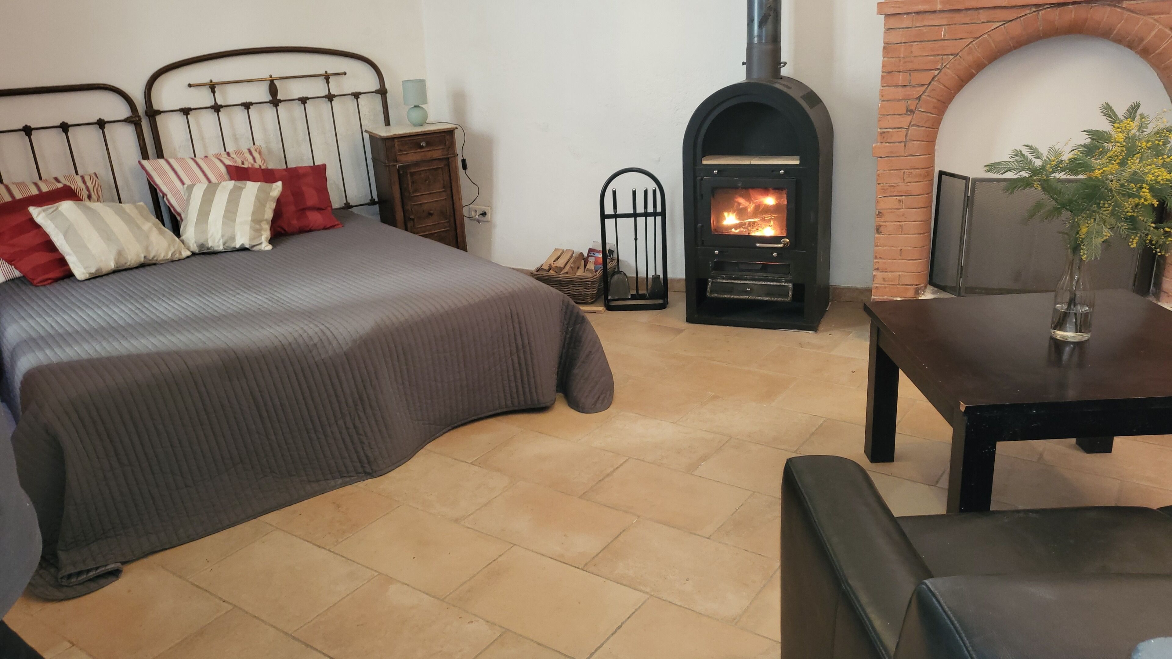 A bedroom with a bed, a nightstand, a wooden table, and a brick fireplace.