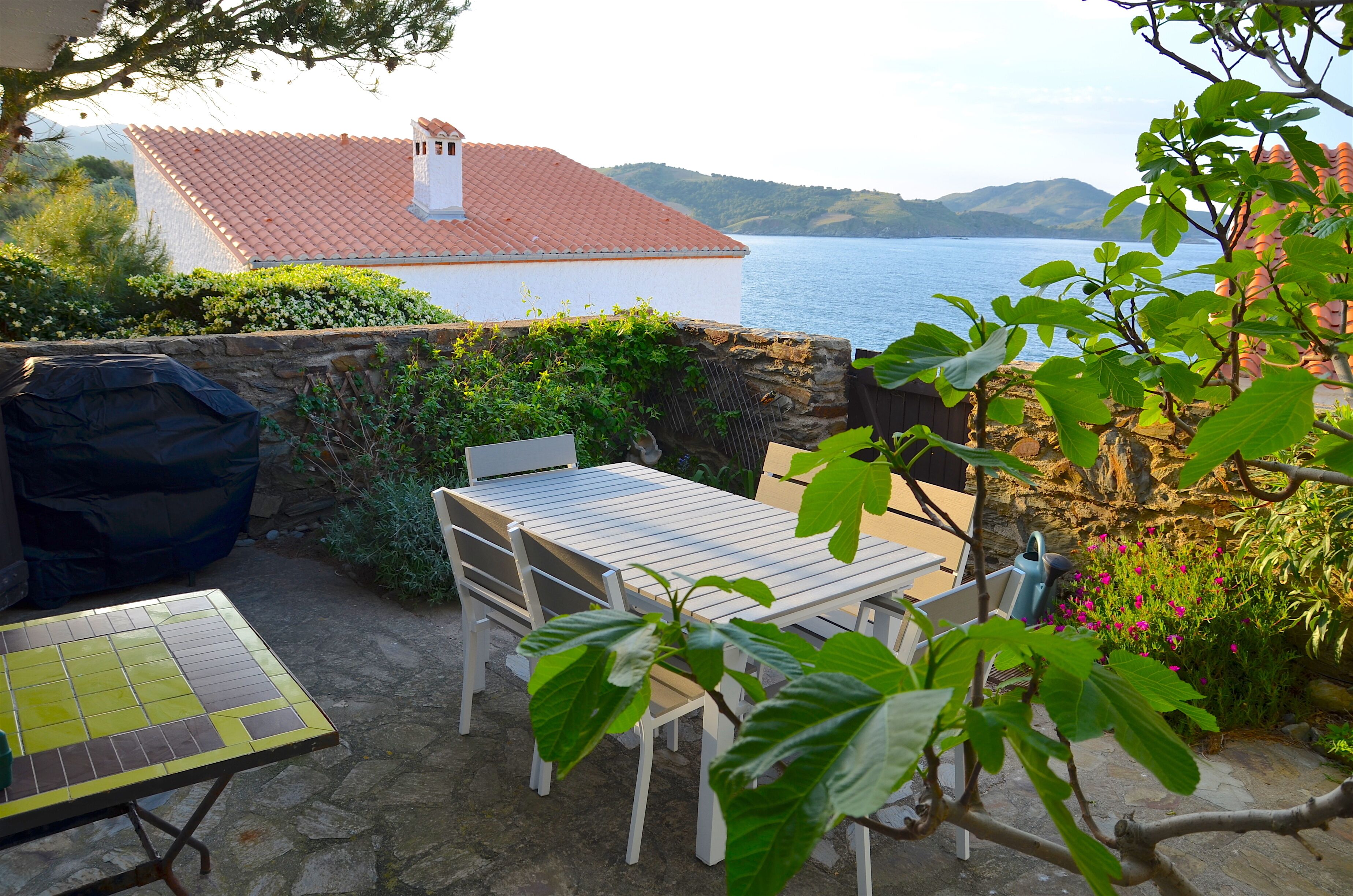 A patio with a table and chairs, a barbecue grill, and a view of the sea and hills.