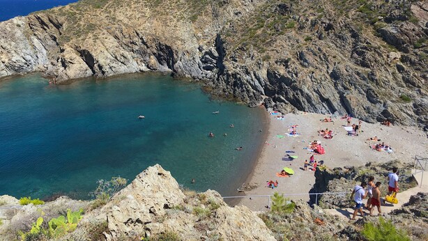 A rocky beach with clear blue water, a sandy area with umbrellas, and a rugged hillside.