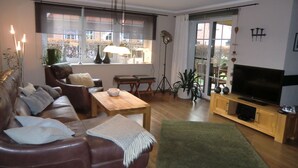 Ein Wohnzimmer mit einer Ledersofa, einem Holztisch, einem Fernseher auf einem Holzständer und einem großen Fenster mit Vorhängen.