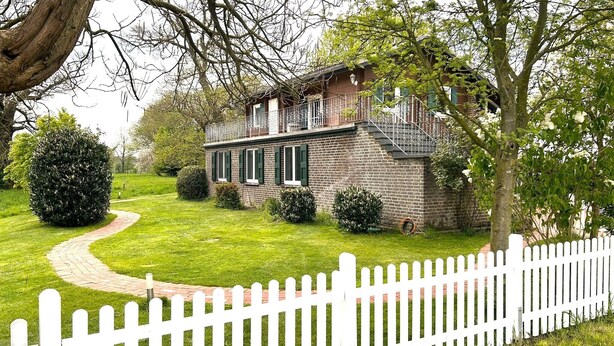A two-story stone house with a brick facade, a balcony with a metal railing, and a white picket fence.