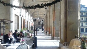 Un café en plein air avec des tables et des chaises, une allée couverte et une vue sur les bâtiments en arrière-plan.