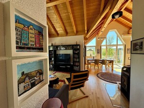 Ein geräumiges Wohnzimmer mit Holzbalkendecke und großen Fenstern, einem Kamin, einem Fernseher und einem Essbereich mit Tisch und Stühlen.