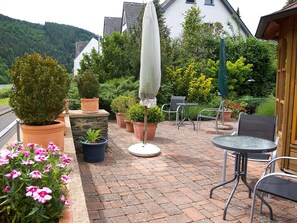 Eine Terrasse mit rundem Tisch und Stühlen, umgeben von Topfpflanzen und einem großen Sonnenschirm.