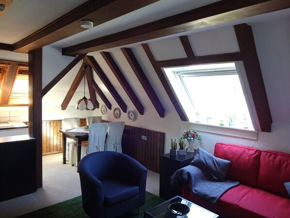Ein gemütliches Wohnzimmer mit einem roten Sofa, einem blauen Sessel und einem Essbereich mit Holzbalken und einem Fenster.