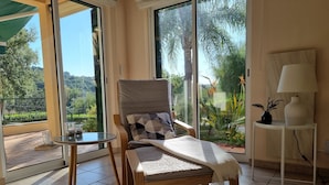 A sunlit room with a wooden chair and a small table, a large window with a view of greenery, and a white lamp on a side table. - Oasis of peace in the Algarve, private pool, heating (Sao Bras de Alportel)