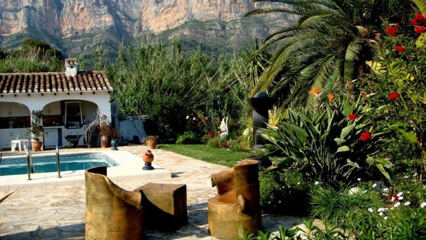 Area piscina con montagne sullo sfondo, circondata da vegetazione lussureggiante e un edificio bianco con tetto in tegole.