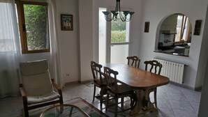 Zona pranzo con tavolo e sedie in legno, un lampadario, una finestra con tende e una cucina visibile attraverso un arco.