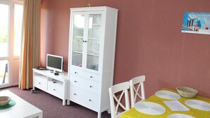 Una habitació amb un armari blanc, una televisió sobre un suport blanc, una taula de dinar amb estovail blanc i plats blancs, i una finestra amb cortines.