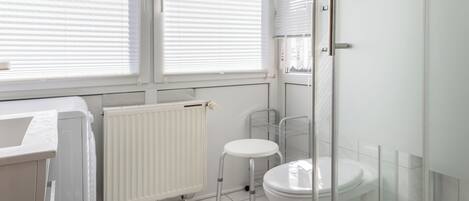 A bathroom with a toilet, a radiator, a window with blinds, and a shower area.