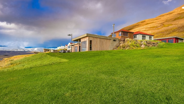 A modern building with large windows and a flat roof, situated on a grassy hillside with a view of a mountain range. - Loft house with incredible views of the fjord (Akureyri)