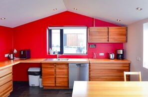 A compact kitchen with a red accent wall, a window, wooden cabinets, a dishwasher, and a coffee maker.