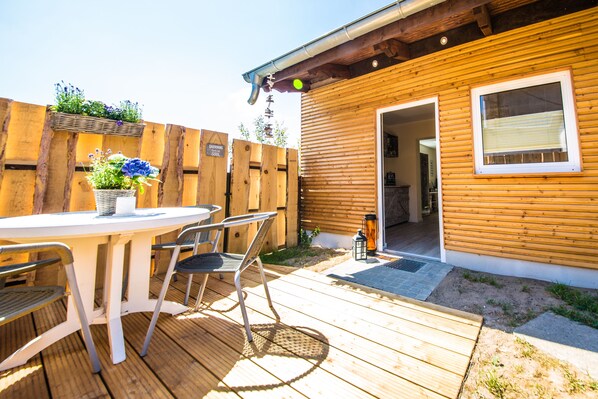 Una terraza de madera con una mesa redonda y sillas, una valla de madera, y un edificio con una ventana y una puerta.