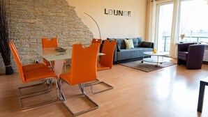 A modern lounge with a stone wall, orange chairs, a glass table, a sofa, and a view through large windows.