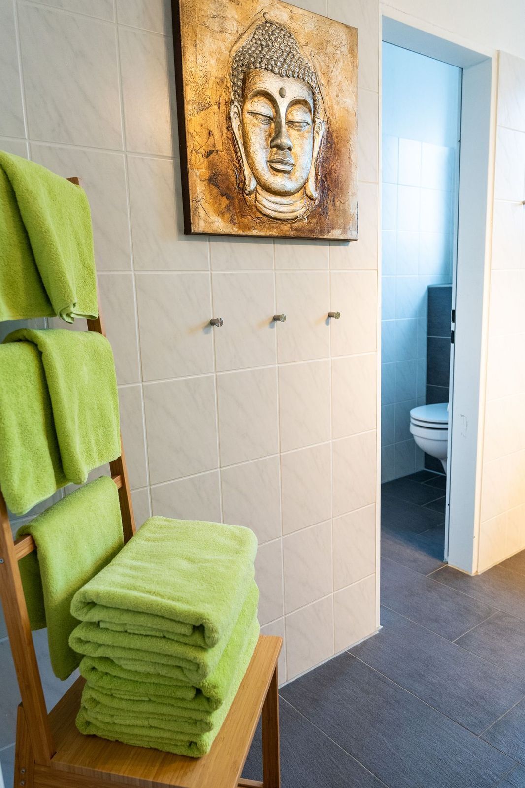 Ein Badezimmer mit einem hölzernen Handtuchhalter, der grüne Handtücher trägt, einem Buddha-Gemälde an der Wand und einer Toilettentür, die offen steht.